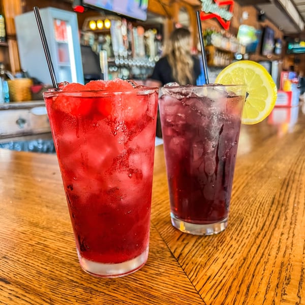 Two vibrant Marquarts Landing cocktails, one red and one purple with a lemon slice, on a bar.