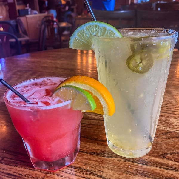 Two cocktails, one red and one yellow with lime and orange slices, on a wooden table.